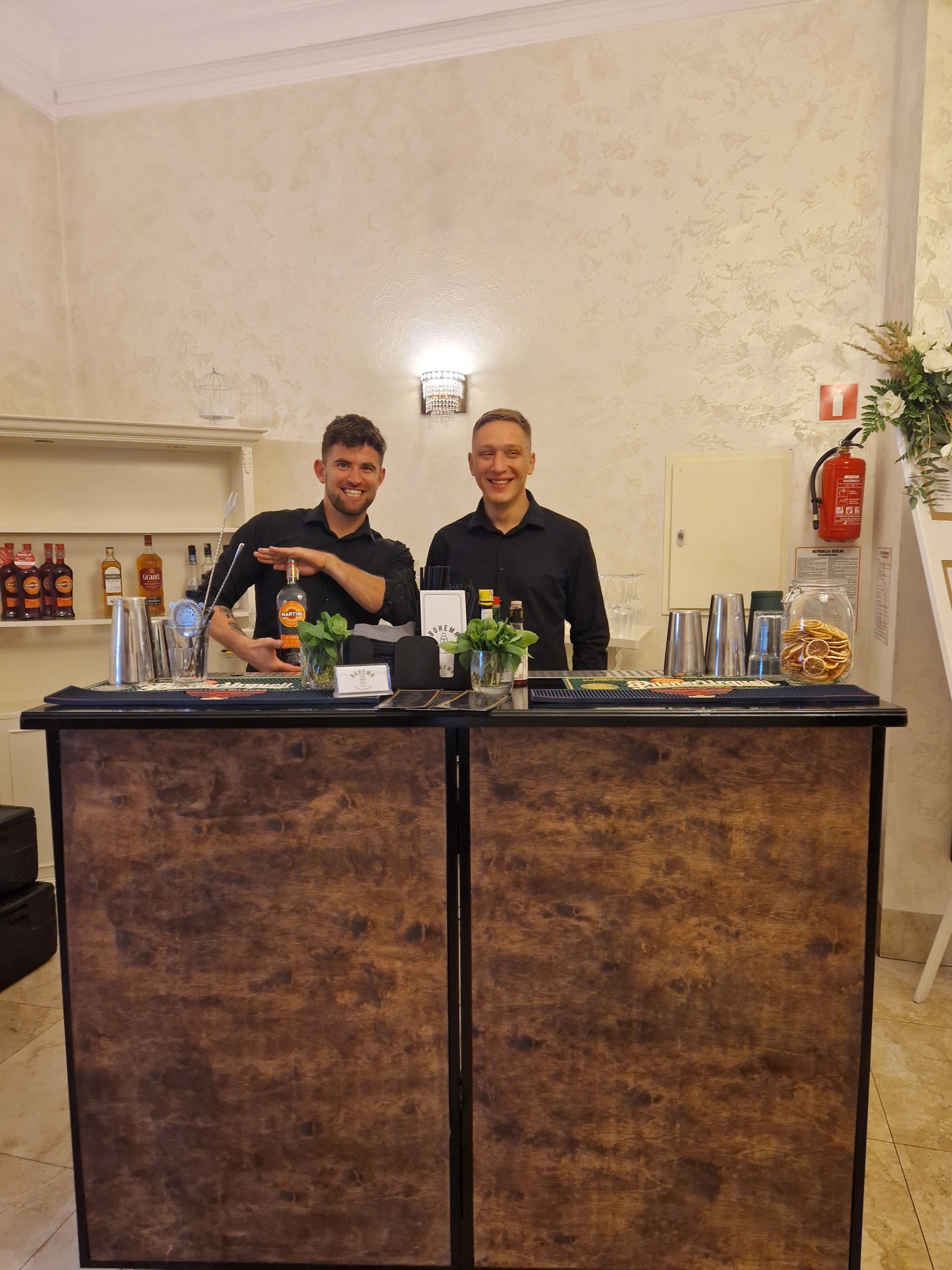 Dwóch uśmiechniętych barmanów za mobilnym barem z drewnianym frontem, z wyposażeniem do przygotowywania drinków i butelką Martini w dłoni jednego z nich.
