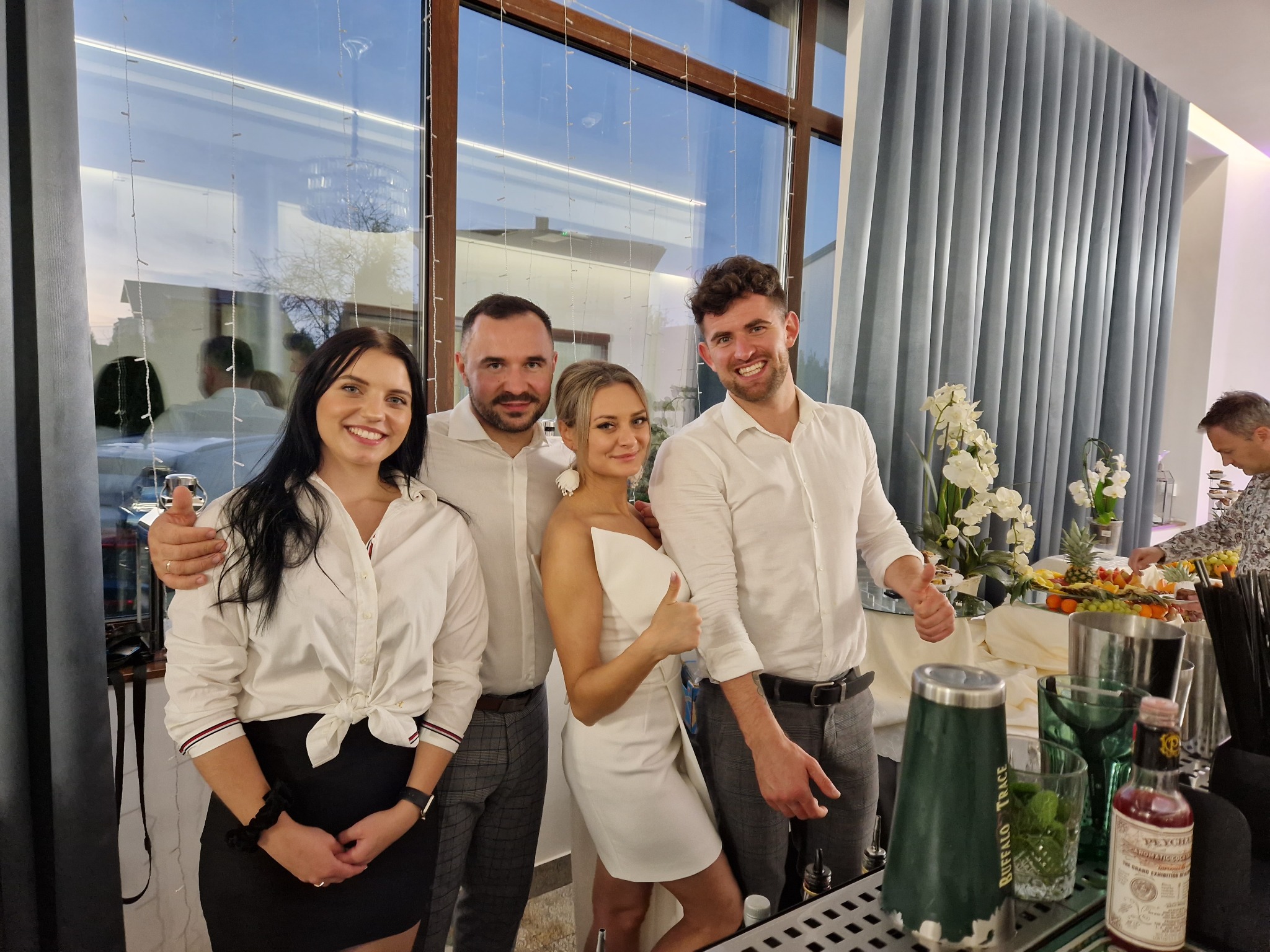 Zespół barmanów w eleganckich strojach pozuje przy barze z widocznymi butelkami Buffalo Trace i Peychaud's Bitters, przygotowującym owoce i kwiatowe dekoracje w tle.