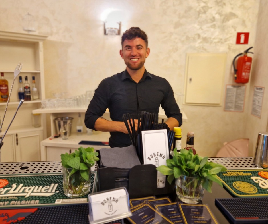 Uśmiechnięty barman za barem z zielonymi dekoracjami z mięty, kartami menu i logiem Bohema, w tle białe szafki i gaśnica.