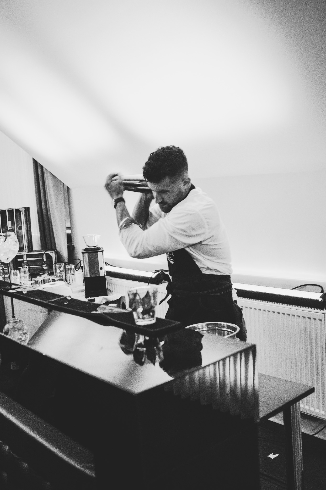 Czarno-białe zdjęcie barmana w białej koszuli i czarnym fartuchu, energicznie mieszającego drinka w shakerze za czarnym barem, z widocznymi szklankami i akcesoriami barmańskimi w tle.