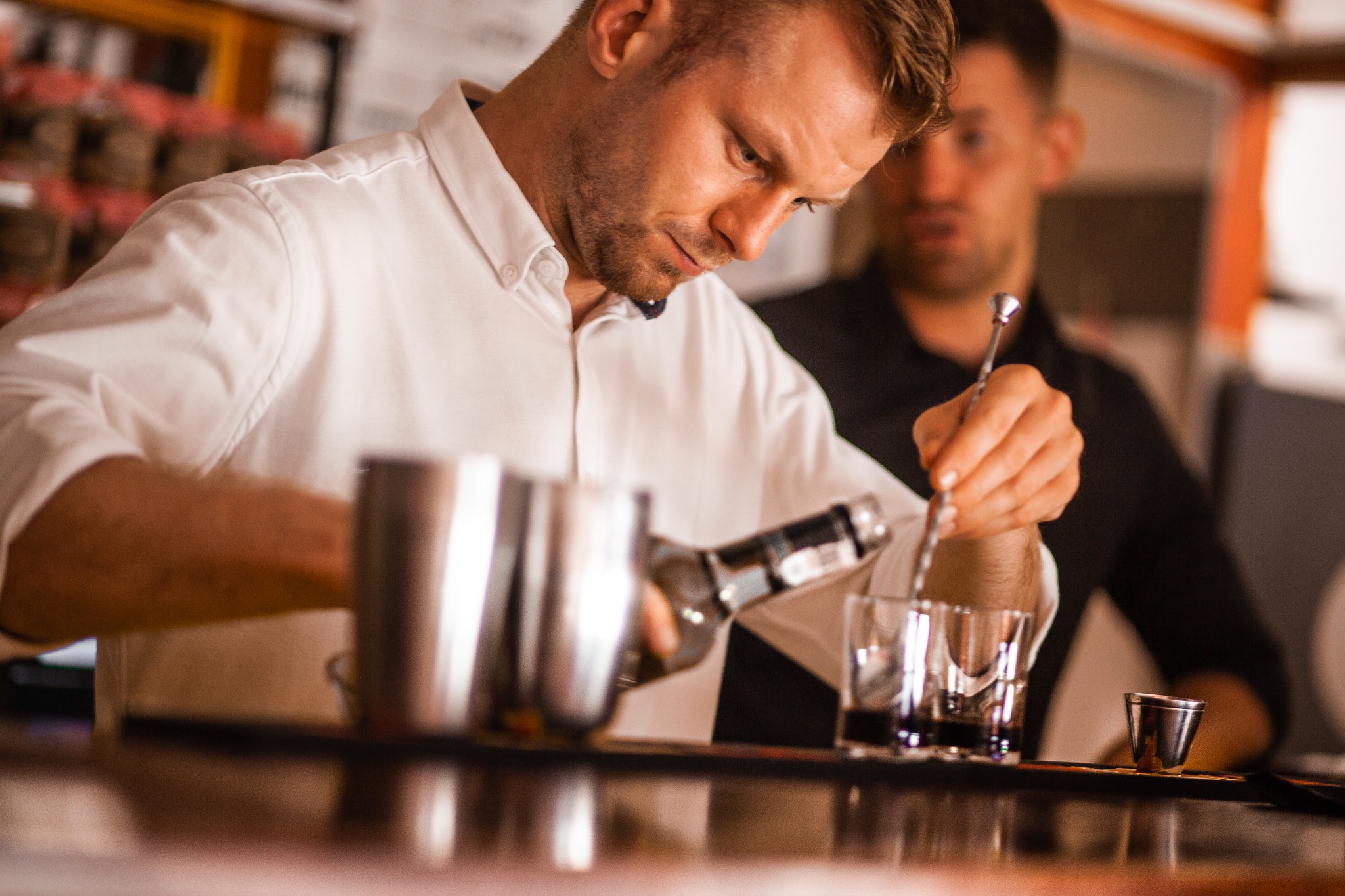 Barman w białej koszuli nalewa ciemny płyn z butelki do kieliszka, obok metalowy shaker i drugi barman mieszający drinka, oświetlenie baru.