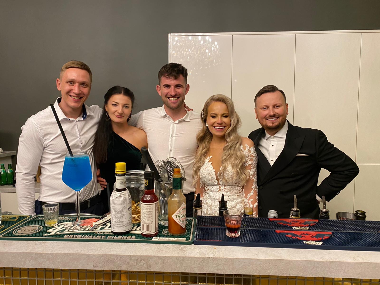 Uśmiechnięci barmani i para młoda za barem z drinkami, w tle białe szafki i szara ściana.