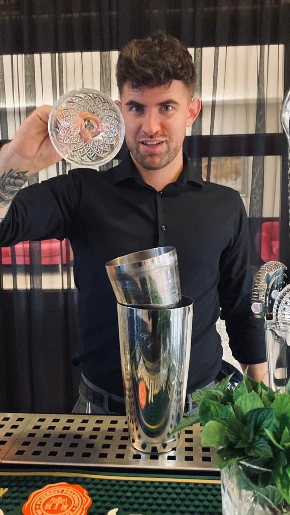 Uśmiechnięty barman w czarnej koszuli prezentuje kryształowy kieliszek nad metalowym shakerem i świeżą miętą na barze.