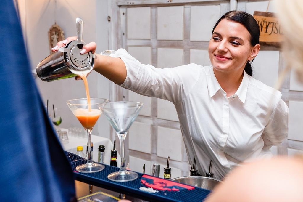 Uśmiechnięta barmanka w białej koszuli przelewa pomarańczowy koktajl ze srebrnego shakera do kieliszka martini, za barem widoczne butelki z alkoholami i akcesoria barmańskie.