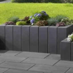 Kompozycja trzech podwyższonych rabat kwiatowych z szarej kostki brukowej, wypełnionych różnorodną zielenią i kwitnącymi roślinami, na tle zielonego trawnika.