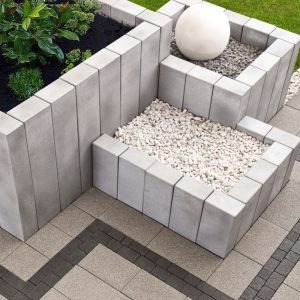 Kaskadowa rabata z szarej kostki betonowej, wypełniona białym grysem i roślinami, z dekoracyjną białą kulą, ułożona na wzorzystej nawierzchni z kostki brukowej.