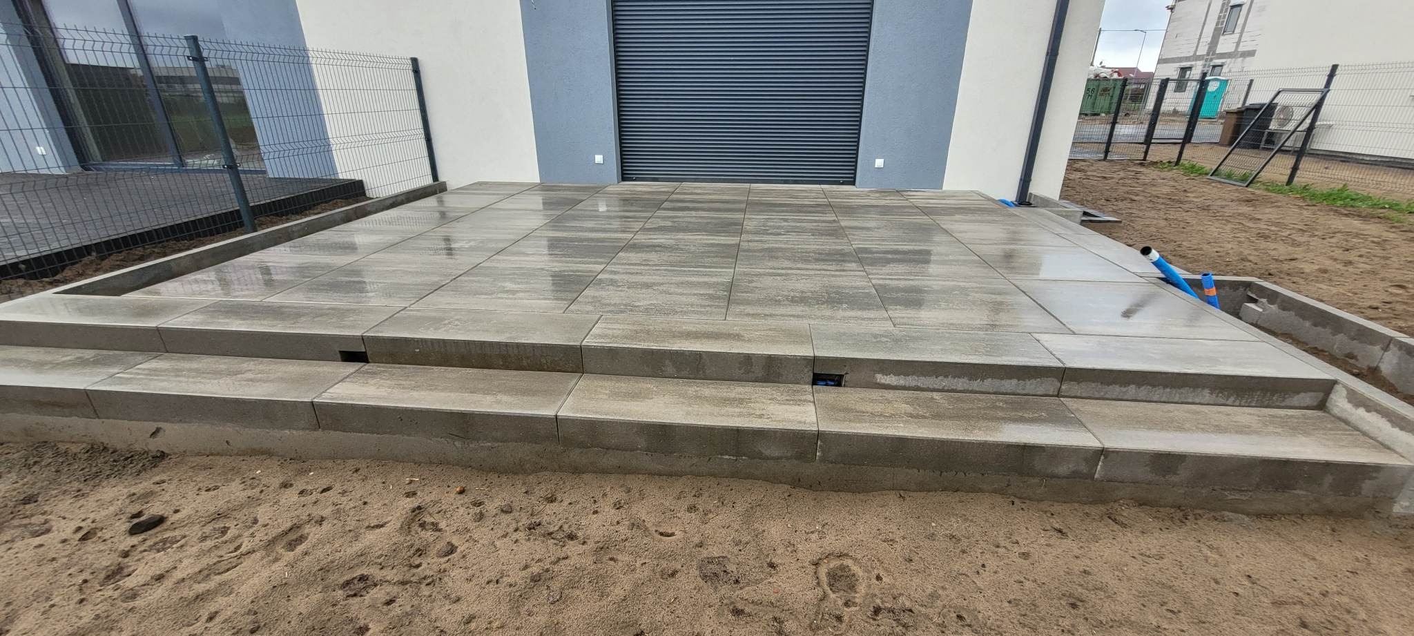 Nowoczesny taras z szarej kostki brukowej ułożonej na dwóch poziomach, z widocznymi niebieskimi rurami wystającymi z boku i fragmentem ogrodzenia panelowego w tle.