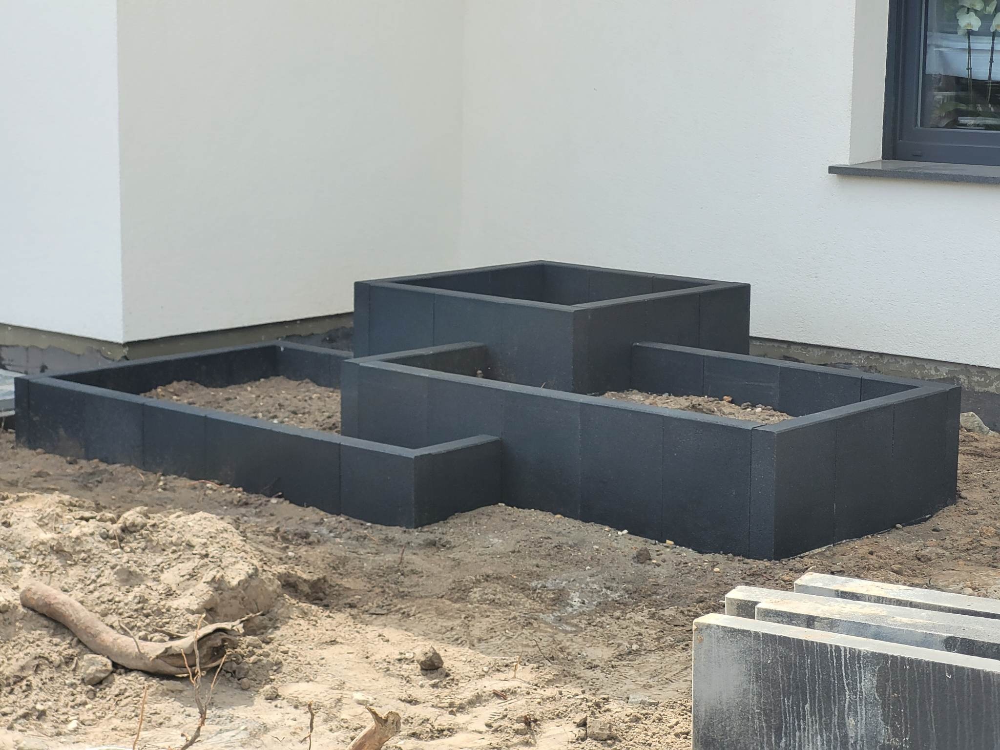 Konstrukcja z szarych bloczków betonowych tworząca podwyższone rabaty na ziemi przy ścianie budynku, widoczne ułożone elementy obok.