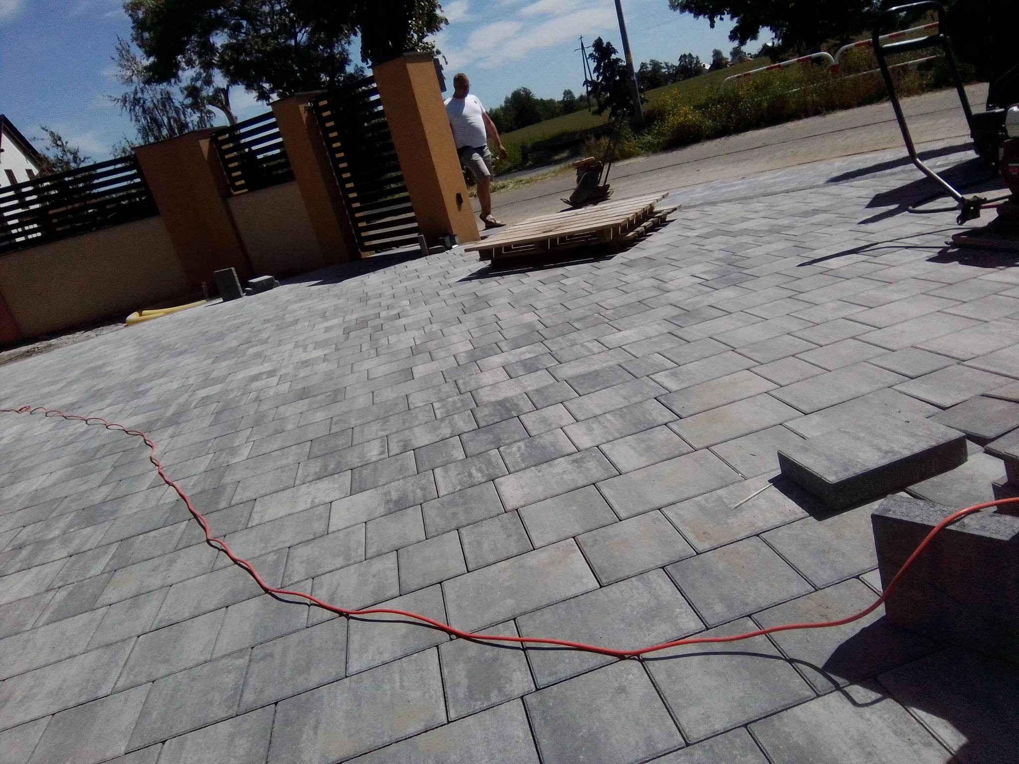 Ułożona szara kostka brukowa na podjeździe, z widocznym fragmentem ogrodzenia, paletą i zagęszczarką w tle, oraz czerwoną kablem zasilającym, ułożonym wzdłuż krawędzi.