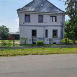 Dwukondygnacyjny dom jednorodzinny z szarą dachówką, jasną elewacją i ciemnymi obramowaniami okien, ogrodzony ozdobnym metalowym płotem na podmurówce z kostki.