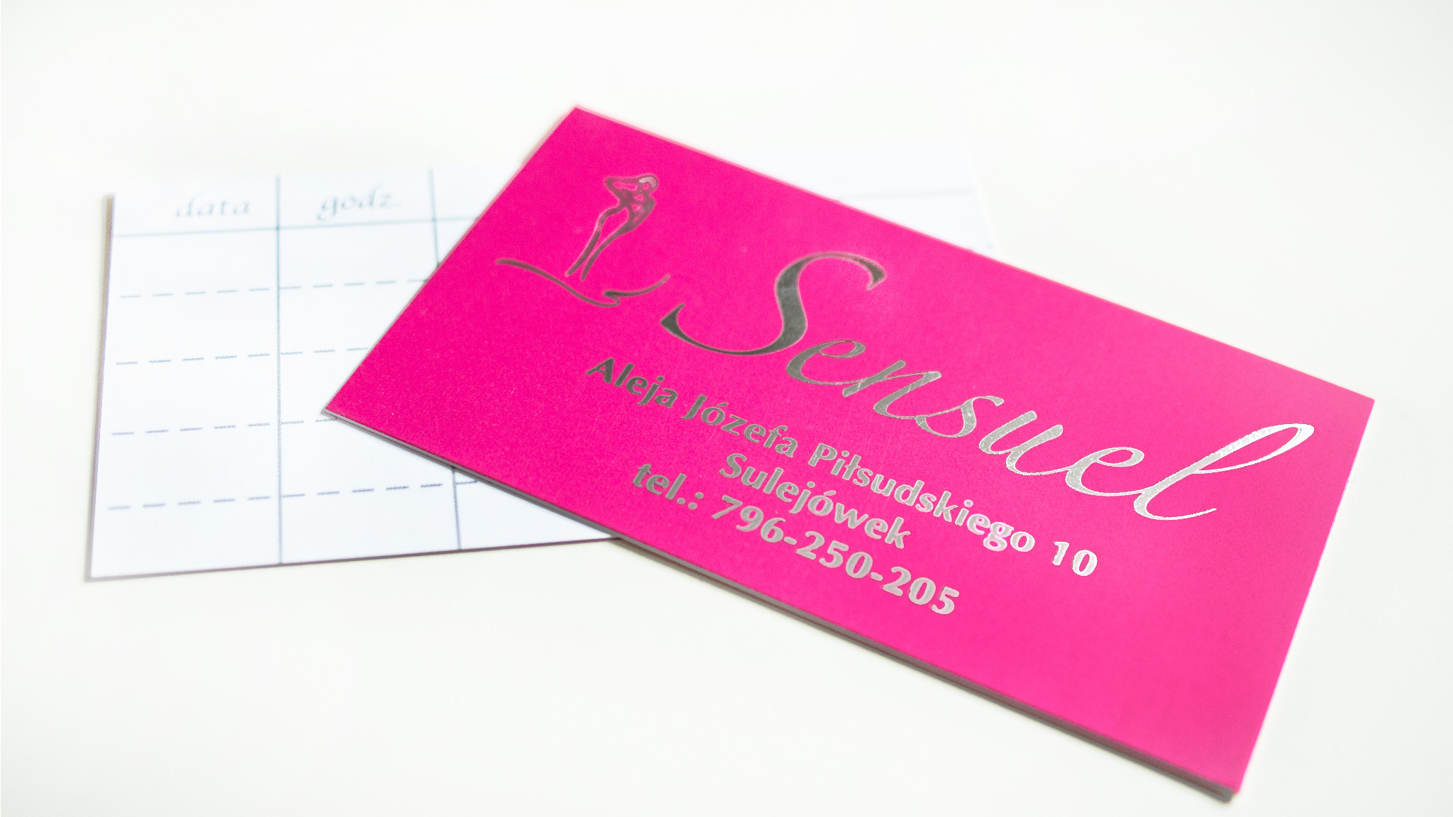 Różowa wizytówka z srebrnym napisem 'Sensuel' i danymi adresowymi, leżąca obok karty z tabelą 'data/godzina' na białym tle.