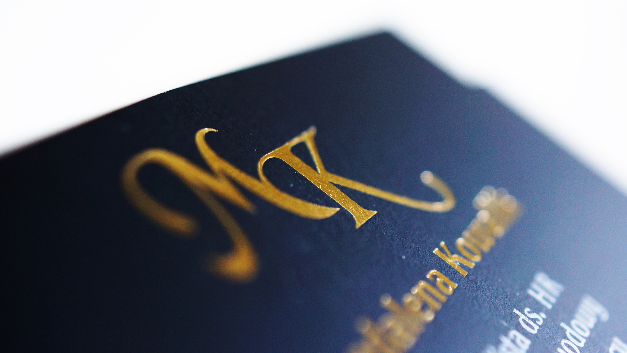 Zbliżenie na wizytówkę z czarnego papieru z wytłoczonym złotym logo i tekstem, widoczne delikatne faktury papieru i refleksy światła.