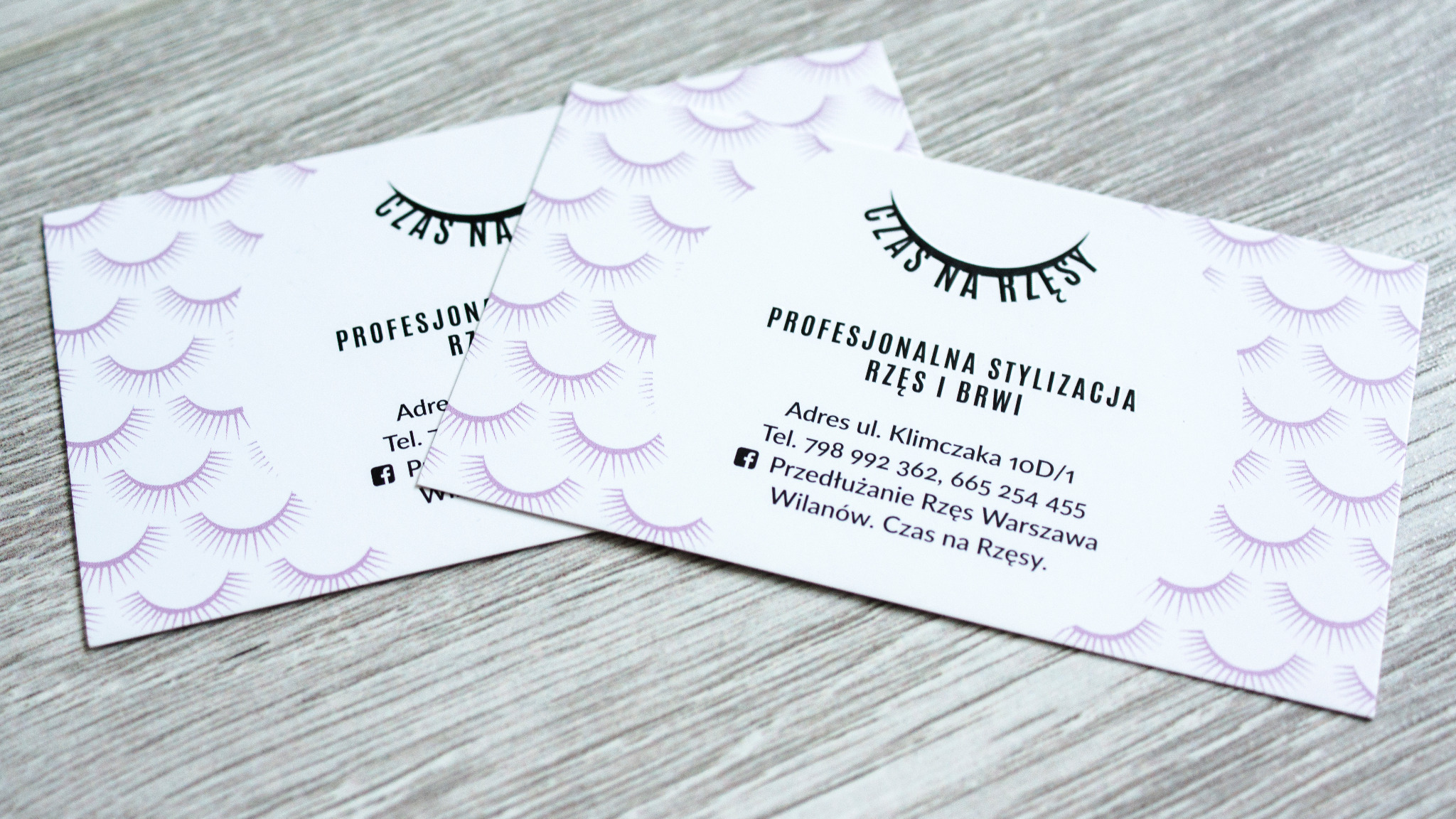 Dwie wizytówki z motywem rzęs i napisem 'Czas na rzęsy', leżące na drewnianym tle.