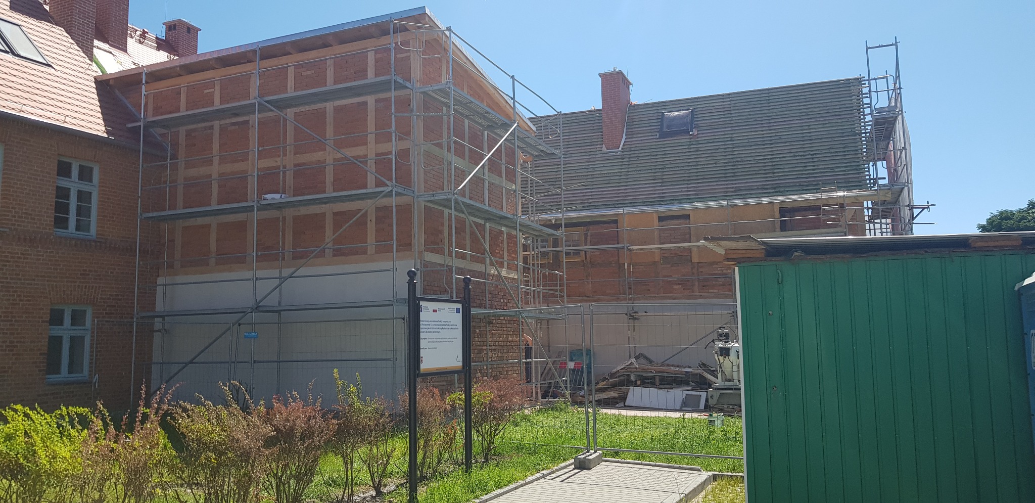 Renowacja ceglanej elewacji budynku z użyciem rusztowań, widoczny fragment nowej izolacji fundamentów i tablica informacyjna