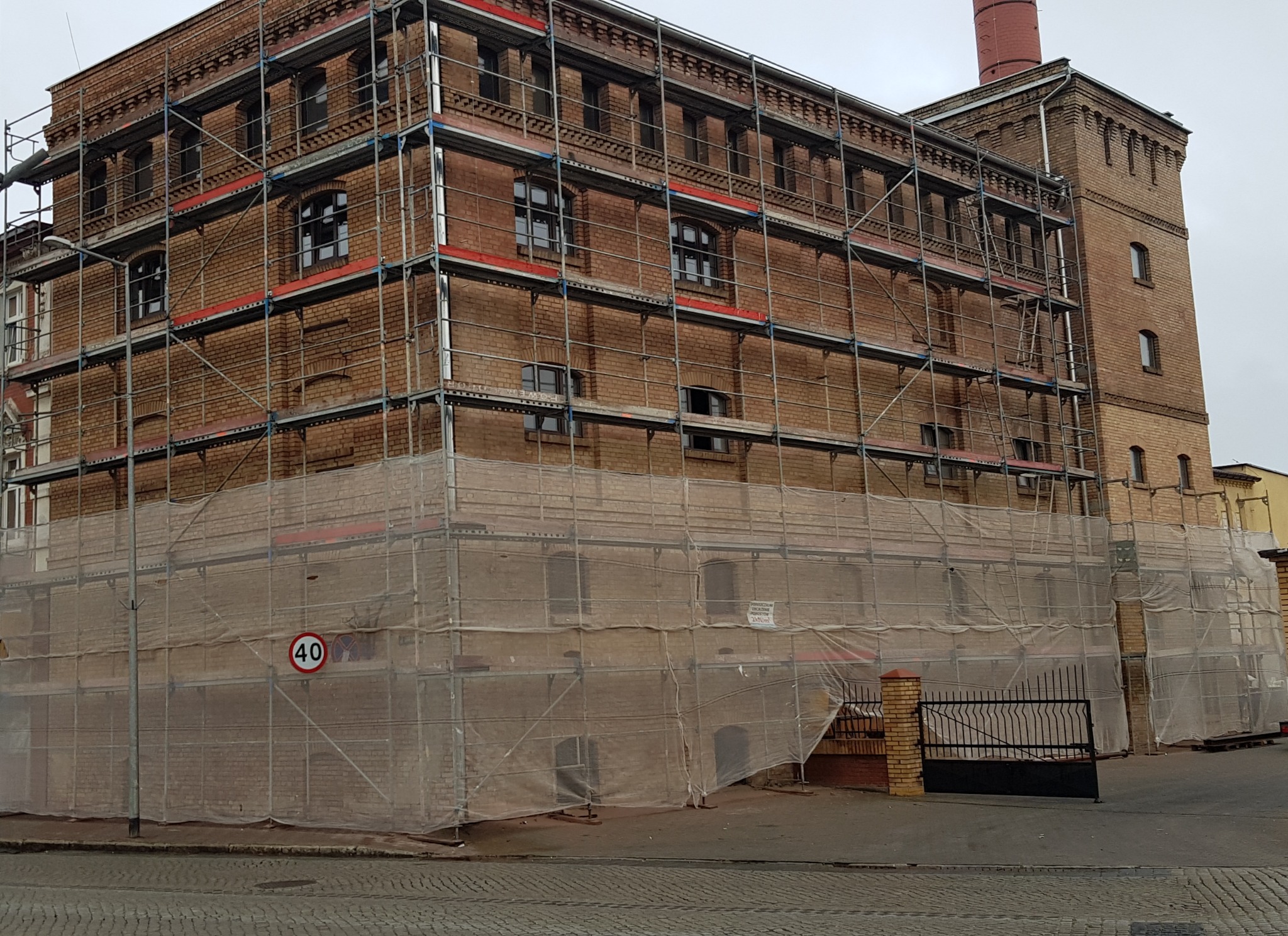 Budynek z cegły w trakcie renowacji, otoczony rusztowaniem i zabezpieczony siatką ochronną, widoczny znak drogowy '40'.