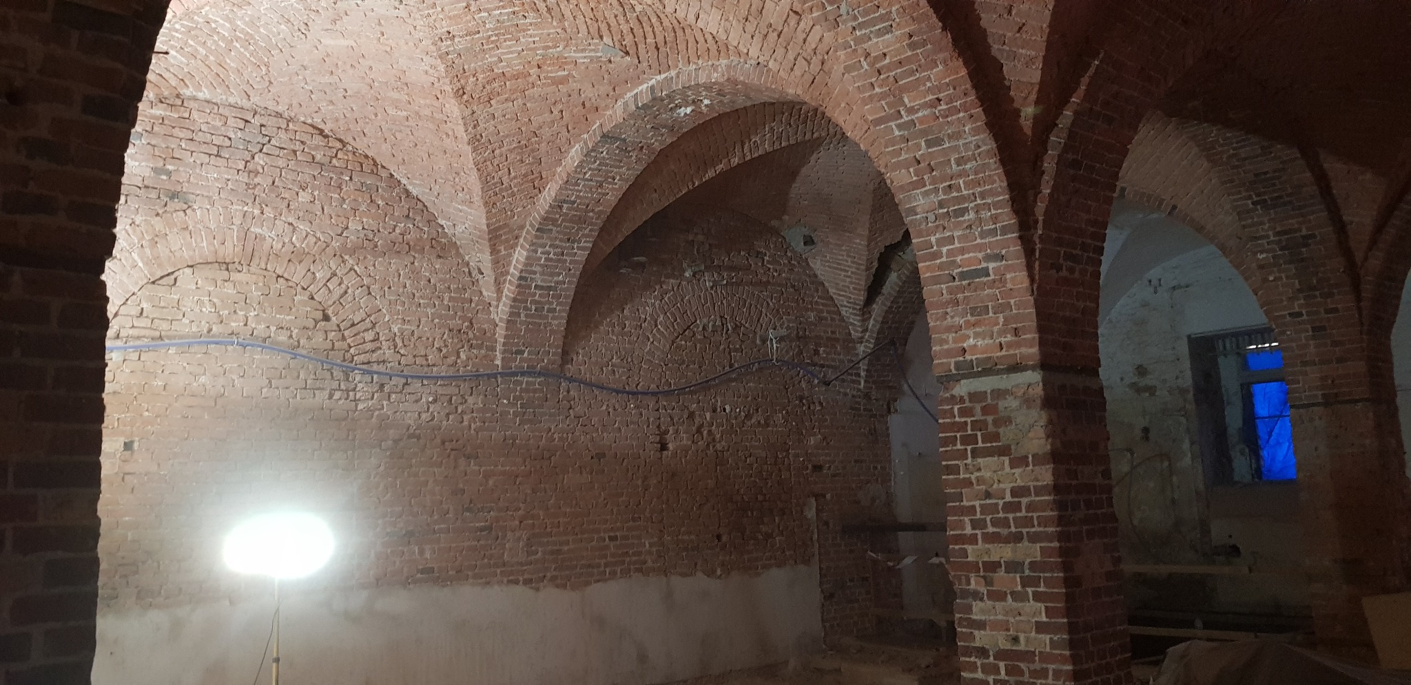 Ceglane sklepienie łukowe w piwnicy lub podziemiach, oświetlone lampą halogenową, z widocznymi śladami wilgoci na ścianach i niebieskim materiałem zasłaniającym okno.