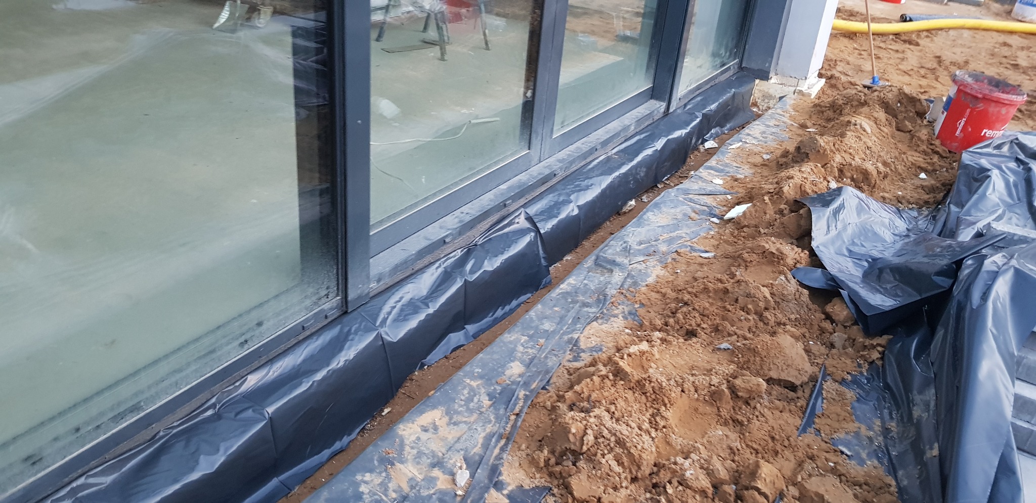 Przygotowanie do izolacji fundamentów: czarna folia ochronna ułożona wzdłuż ściany budynku z dużymi oknami, wykop z ziemią, widoczny czerwony kubeł i narzędzia w tle.