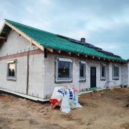 WINDOBEST SP. Z O.O - Niedokończony dom z pustaków z zielonym dachem, oknami z roletami zewnętrznymi i czarnymi drzwiami wejściowymi. Na pierwszym planie worki z zaprawą.