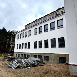 WINDOBEST SP. Z O.O - Nowy budynek z szarymi oknami PCV i stertą ram okiennych na ziemi. Widoczne rusztowania i drzewa w tle. Budowa w toku, pochmurny dzień.