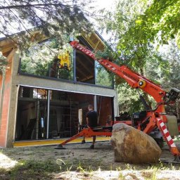 WINDOBEST SP. Z O.O - Montaż okien z użyciem mini żurawia na tle domu w stanie surowym otwartym, otoczonego zielenią. Widoczny pracownik obserwujący proces.