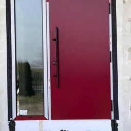 WINDOBEST SP. Z O.O - Czerwone drzwi wejściowe z długim uchwytem i wąskim oknem bocznym, białe ramy, widoczne uszczelnienia i surowy mur, ujęcie z bliska, bez otoczenia.