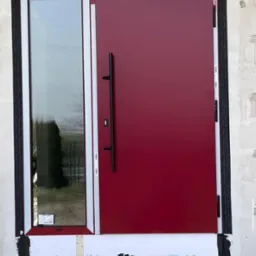 Czerwone drzwi wejściowe z długim uchwytem i wąskim oknem bocznym, białe ramy, widoczne uszczelnienia i surowy mur, ujęcie z bliska, bez otoczenia.