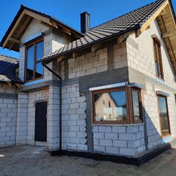 WINDOBEST SP. Z O.O - Dom w trakcie budowy: surowe mury z oknami w brązowych ramach, czarne rynny, dach z ciemnej dachówki, błękitne niebo w tle. Widoczny etap montażu okien.