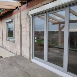 WINDOBEST SP. Z O.O - Szare okna PCV z roletą zewnętrzną w surowym budynku z betonu komórkowego. Widoczne elementy konstrukcji dachu i mniejsze okna. Krajobraz za oknami.