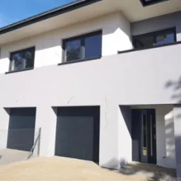 Nowoczesny dom z grafitowymi bramami garażowymi i drzwiami wejściowymi w Jelonku. Minimalistyczna fasada, czarne ramy okienne.