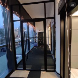 WINDOBEST SP. Z O.O - Nowoczesne, czarne drzwi wewnętrzne z przeszkleniami, oddzielające przestrzeń biurową. Widoczne oświetlenie dekoracyjne i fragment okna z widokiem na zewnątrz.