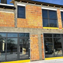 WINDOBEST SP. Z O.O - Surowy stan budynku z zamontowanymi oknami i drzwiami z czarnymi ramami. Widoczny mur z pustaków i ocieplenie. Zimowy krajobraz w tle.