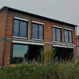 WINDOBEST SP. Z O.O - Surowy budynek z oknami i roletami zewnętrznymi na dwóch kondygnacjach, widok z zewnątrz na tle zieleni. Częściowo opuszczone rolety.