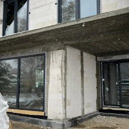 WINDOBEST SP. Z O.O - Surowy stan domu w zimowej scenerii: widoczne okna z czarnymi ramami, białe ściany z betonu komórkowego i deski pod śniegiem. Trwa budowa.