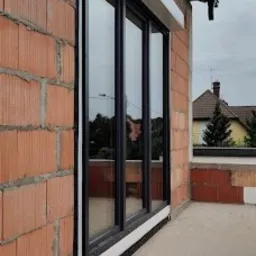 Czarne okno z roletą zewnętrzną na surowej, ceglanej ścianie budynku w trakcie budowy. Widoczne uszczelnienia okienne i fragment dachu.