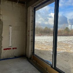 WINDOBEST SP. Z O.O - Nowoczesne okno z czarną ramą w surowym wnętrzu, widok na zaśnieżone pole i drzewa. Instalacja elektryczna widoczna na ścianie.