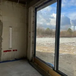 Nowoczesne okno z czarną ramą w surowym wnętrzu, widok na zaśnieżone pole i drzewa. Instalacja elektryczna widoczna na ścianie.