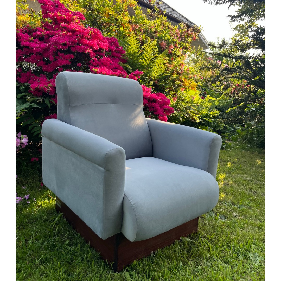 Szary fotel obrotowy z drewnianą podstawą na trawie, w tle kwitnące krzewy różowe i zielone