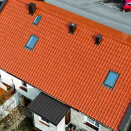 Widok z góry na dom z nową, pomarańczową dachówką ceramiczną, trzema kominami i czterema oknami dachowymi, obok zaparkowany czerwony samochód.