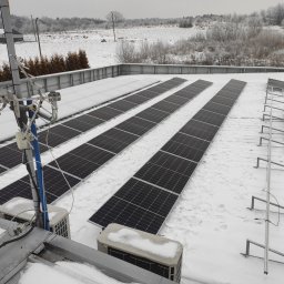 ELPRO Piotr Pisaniak - Instalacja paneli fotowoltaicznych na dachu budynku pokrytym śniegiem, widoczne jednostki zewnętrzne klimatyzacji również pod śniegiem, antena na maszcie i zimowy krajobraz w tle.