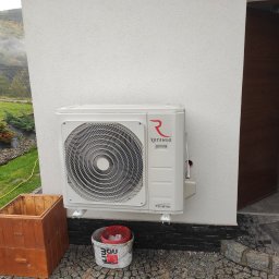 ELPRO Piotr Pisaniak - Biała jednostka zewnętrzna klimatyzacji ROTENSO zamontowana na elewacji domu, obok drewniana skrzynia i wiadro po farbie, widok na zewnątrz z otwartymi drzwiami do pomieszczenia gospodarczego.