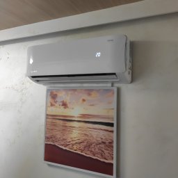 ELPRO Piotr Pisaniak - Biały klimatyzator z wyświetlaczem temperatury (20 stopni) zamontowany nad obrazem z motywem plaży w Rzeszowie, na ścianie o delikatnej fakturze.