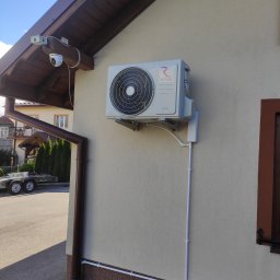 ELPRO Piotr Pisaniak - Zewnętrzna jednostka klimatyzacji Rotenso zamontowana na ścianie domu z widocznymi rurami odprowadzającymi skropliny i przewodami elektrycznymi w białych listwach, z kamerą monitoringu pod okapem...