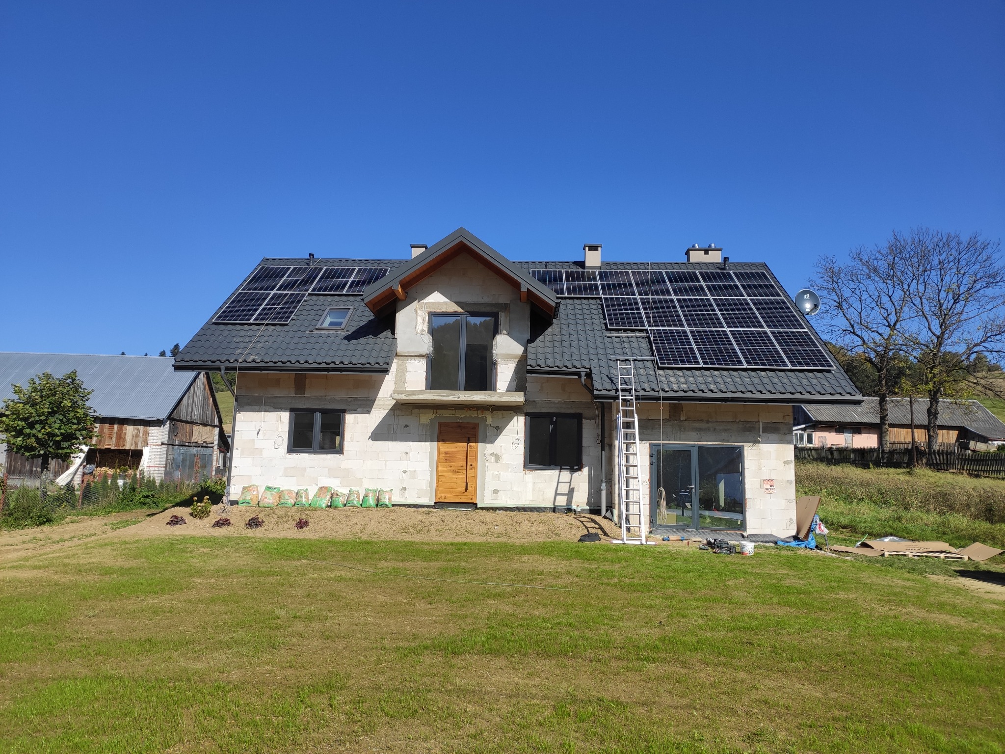 Nowy dom w trakcie budowy z zainstalowanymi panelami słonecznymi na dachu, widoczne worki z materiałami budowlanymi i drabina oparta o ścianę.