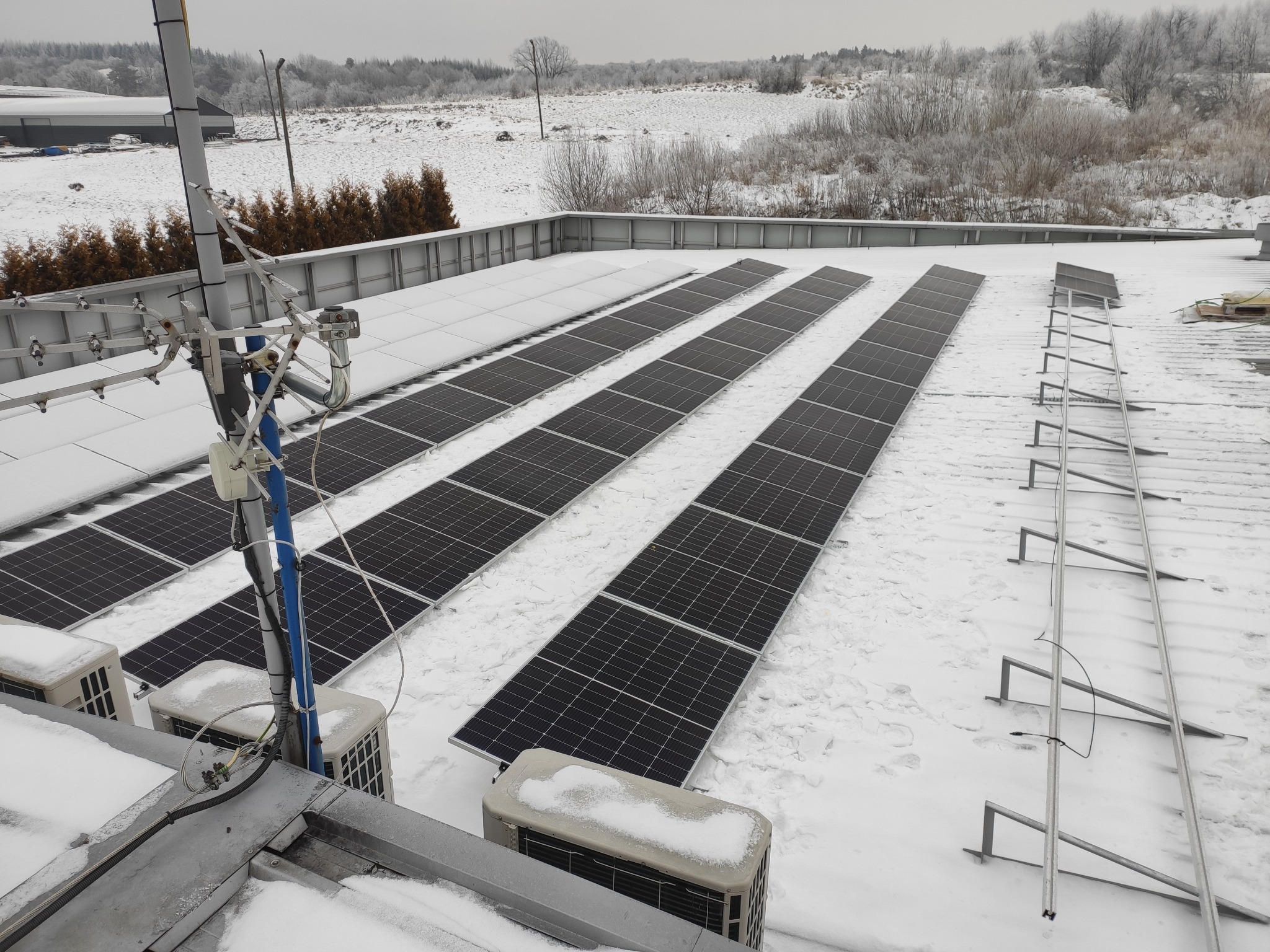 Instalacja paneli fotowoltaicznych na dachu budynku pokrytym śniegiem, widoczne jednostki zewnętrzne klimatyzacji również pod śniegiem, antena na maszcie i zimowy krajobraz w tle.