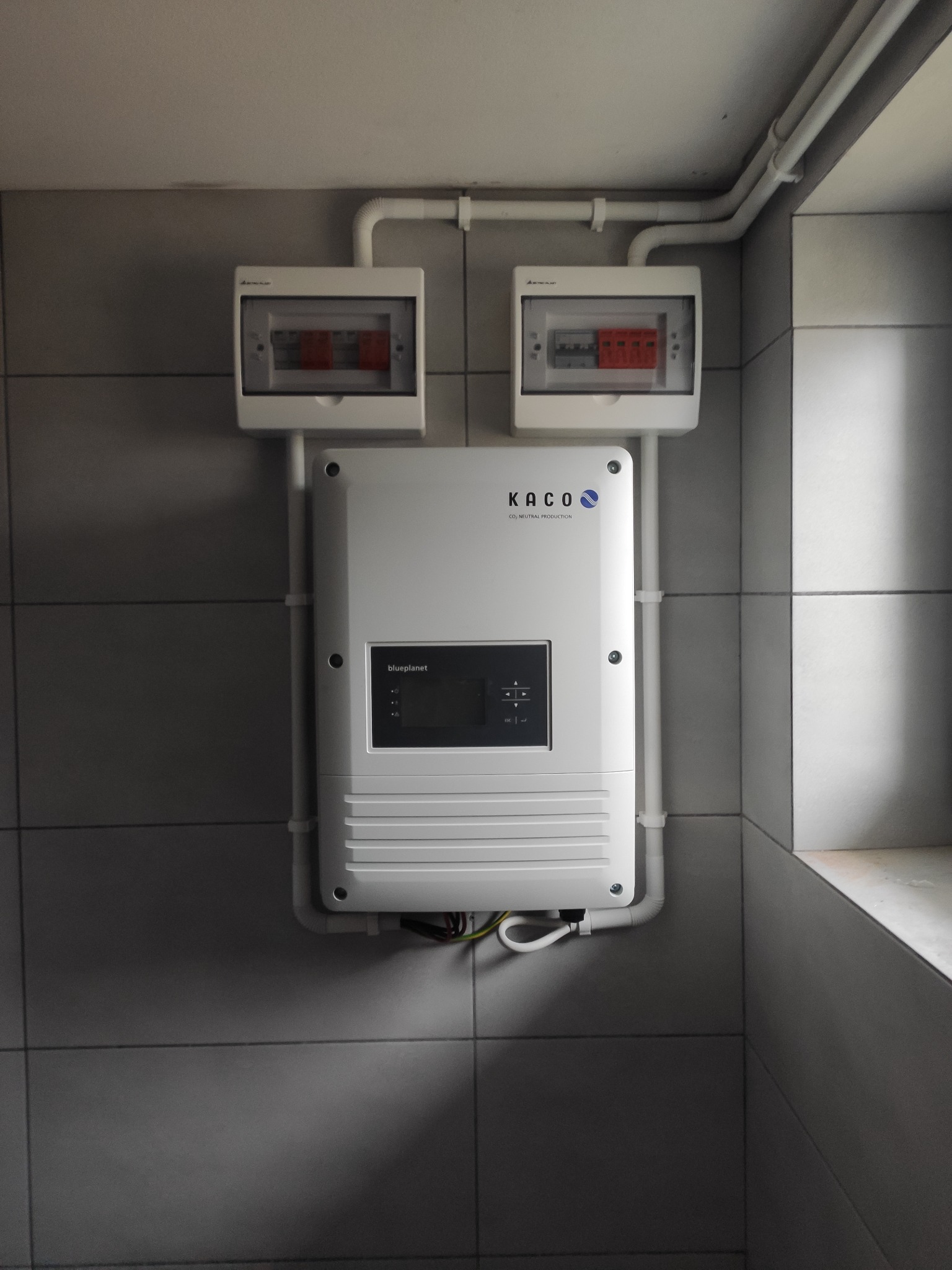 Biała skrzynka z bezpiecznikami i falownik Kaco Blueplanet zamontowane na szarej, kafelkowanej ścianie, z widocznymi przewodami w białych rurkach PCV.
