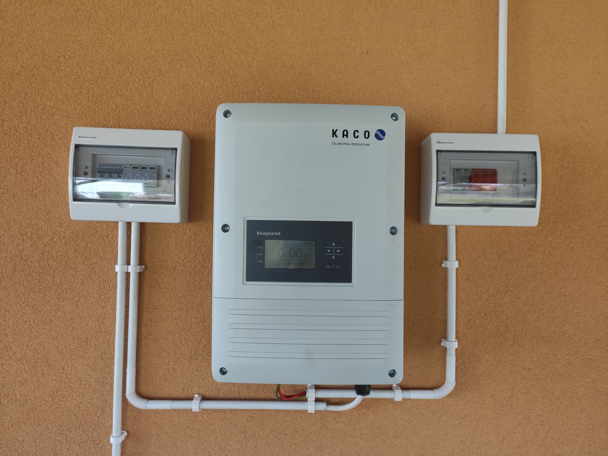 Biała skrzynka elektryczna z wyświetlaczem i logo KACO, zamontowana na pomarańczowej ścianie, z dwiema mniejszymi skrzynkami po bokach i białymi rurami PCV.