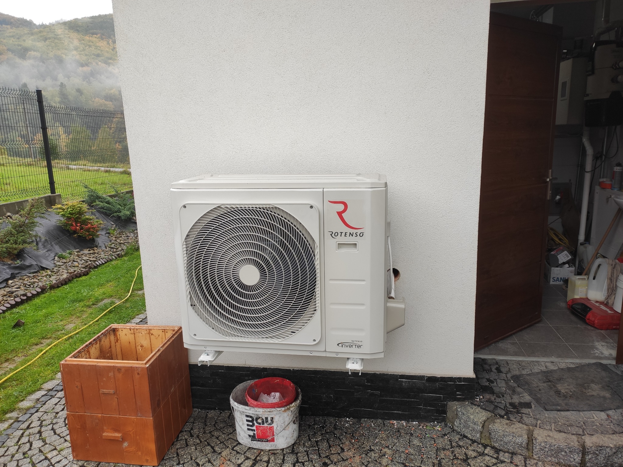 Biała jednostka zewnętrzna klimatyzacji ROTENSO zamontowana na elewacji domu, obok drewniana skrzynia i wiadro po farbie, widok na zewnątrz z otwartymi drzwiami do pomieszczenia gospodarczego.
