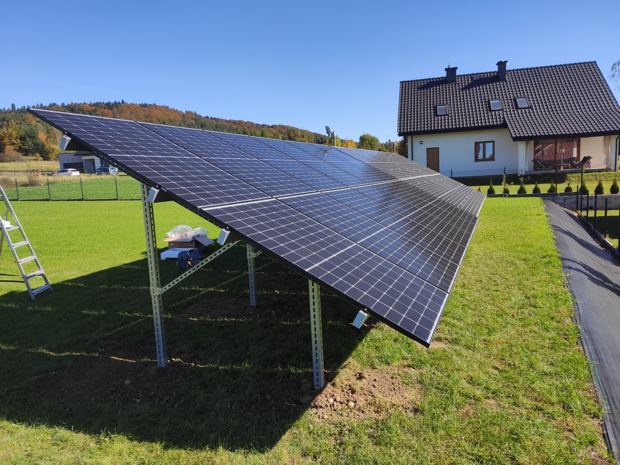 Zestaw paneli fotowoltaicznych na metalowej konstrukcji, świeżo zamontowany na trawniku przed domem jednorodzinnym w słoneczny dzień. Widoczna drabina i kartonowe pudła sugerujące trwający montaż.
