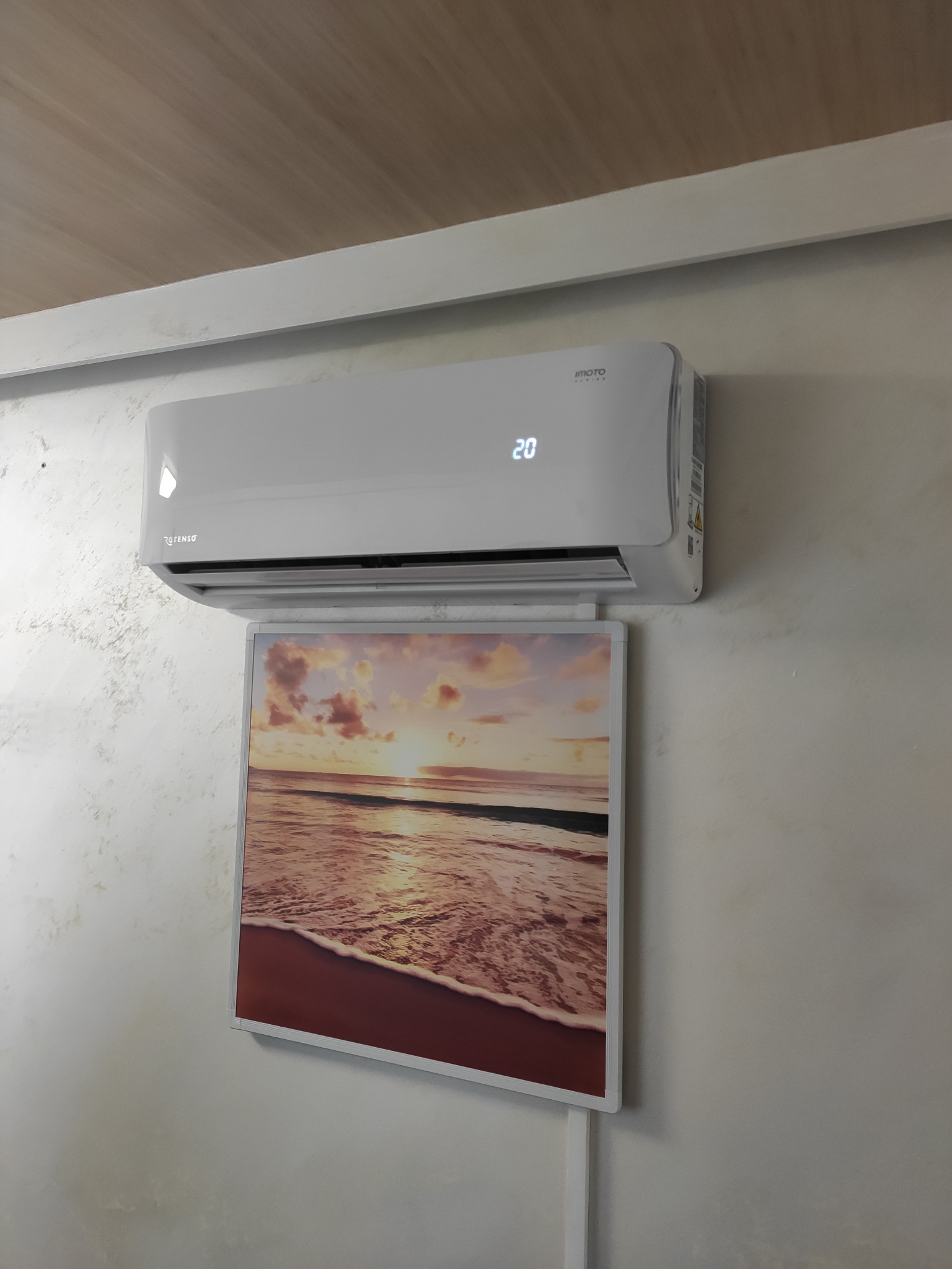 Biały klimatyzator z wyświetlaczem temperatury (20 stopni) zamontowany nad obrazem z motywem plaży w Rzeszowie, na ścianie o delikatnej fakturze.