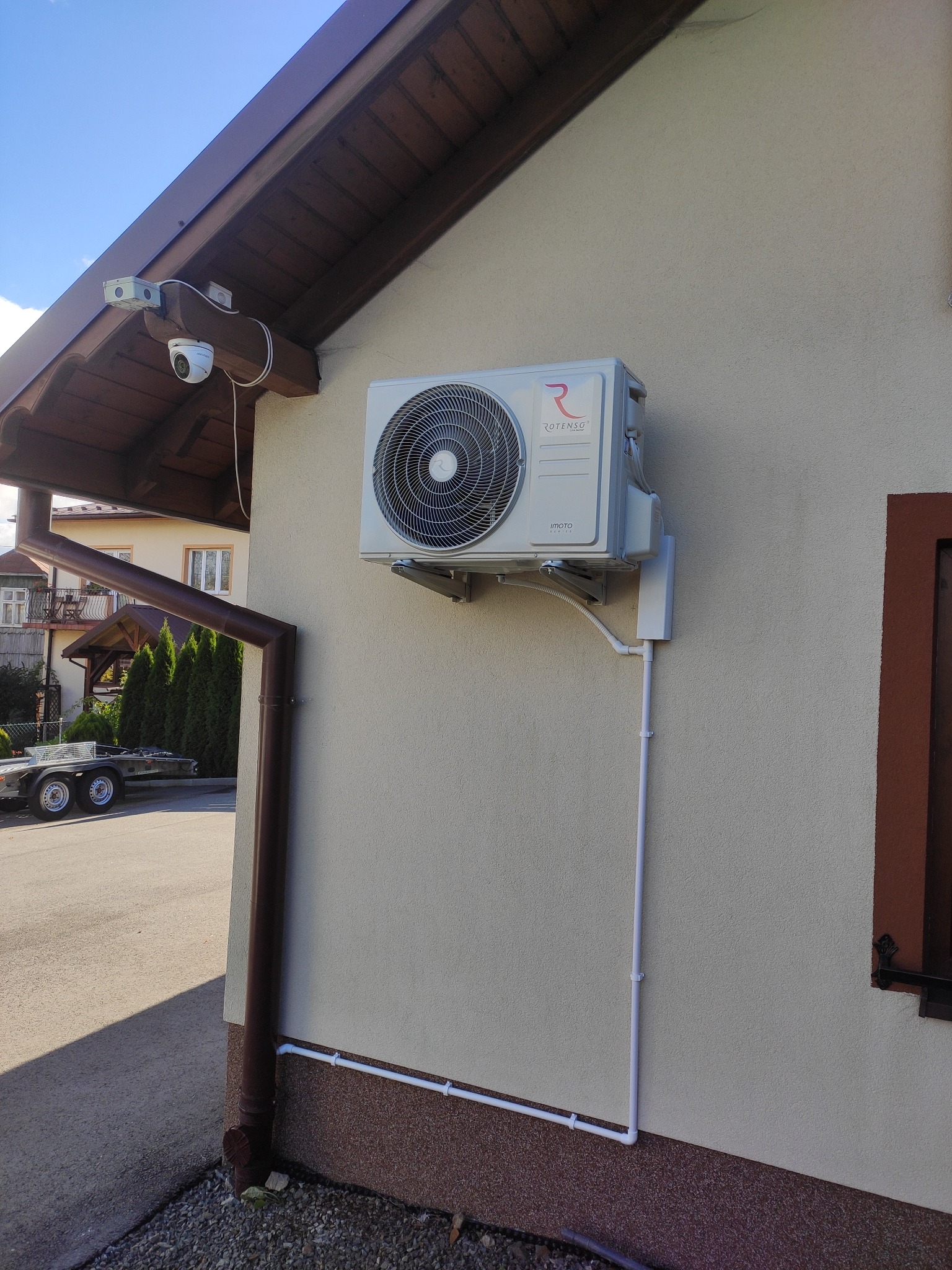 Zewnętrzna jednostka klimatyzacji Rotenso zamontowana na ścianie domu z widocznymi rurami odprowadzającymi skropliny i przewodami elektrycznymi w białych listwach, z kamerą monitoringu pod okapem...